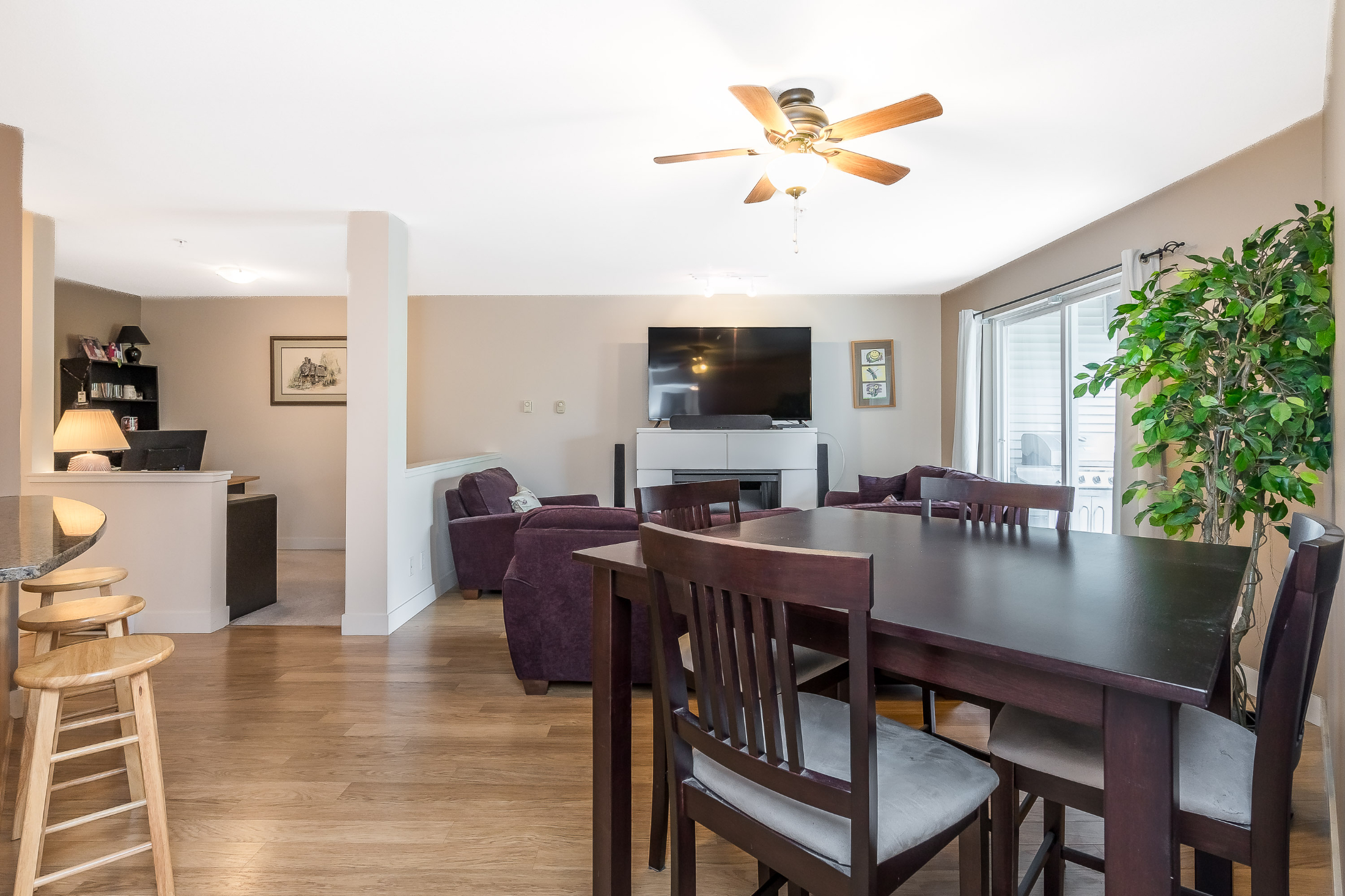 Open living and dining area in Clayton condo featuring wooden dining table, chairs, ceiling fan, and large windows leading to balcony.