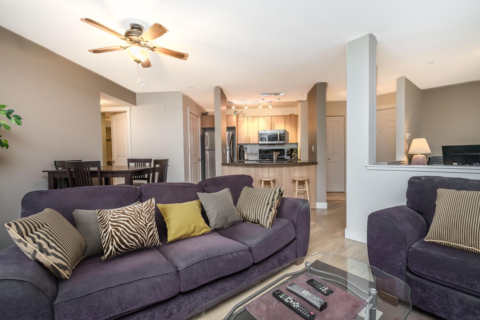 South-facing 1 bed + den condo in Clayton featuring open living room with purple sofa, glass coffee table, and kitchen with stainless steel appliances.